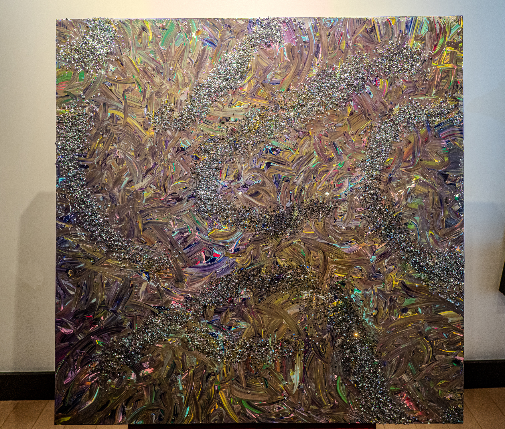Abstract metallic painting with iridescent, swirling brushstrokes and silver glitter trails across a tall canvas in a gallery setting