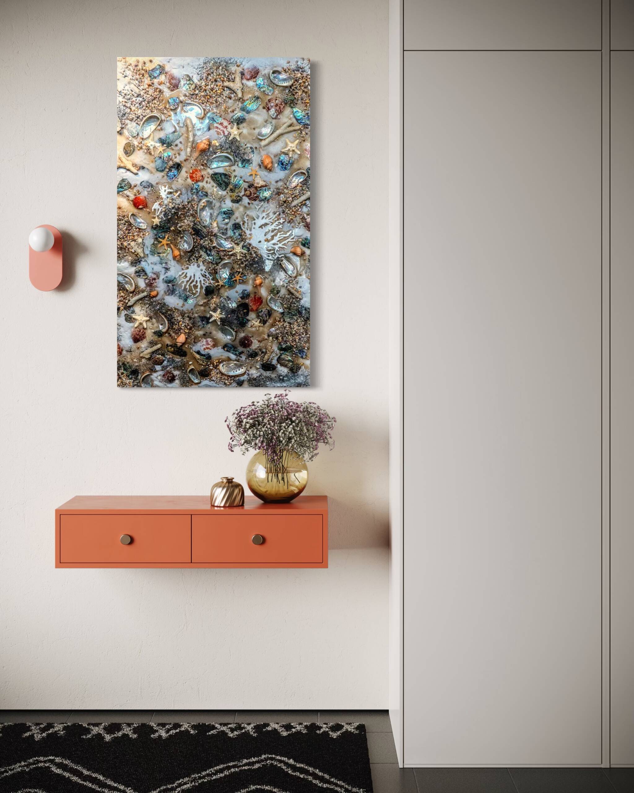 Coral-pink wall shelf with two drawers sits below a colorful sea-shell art piece and a vase of flowers on a peach cabinet, against a light wall and gray built-ins.