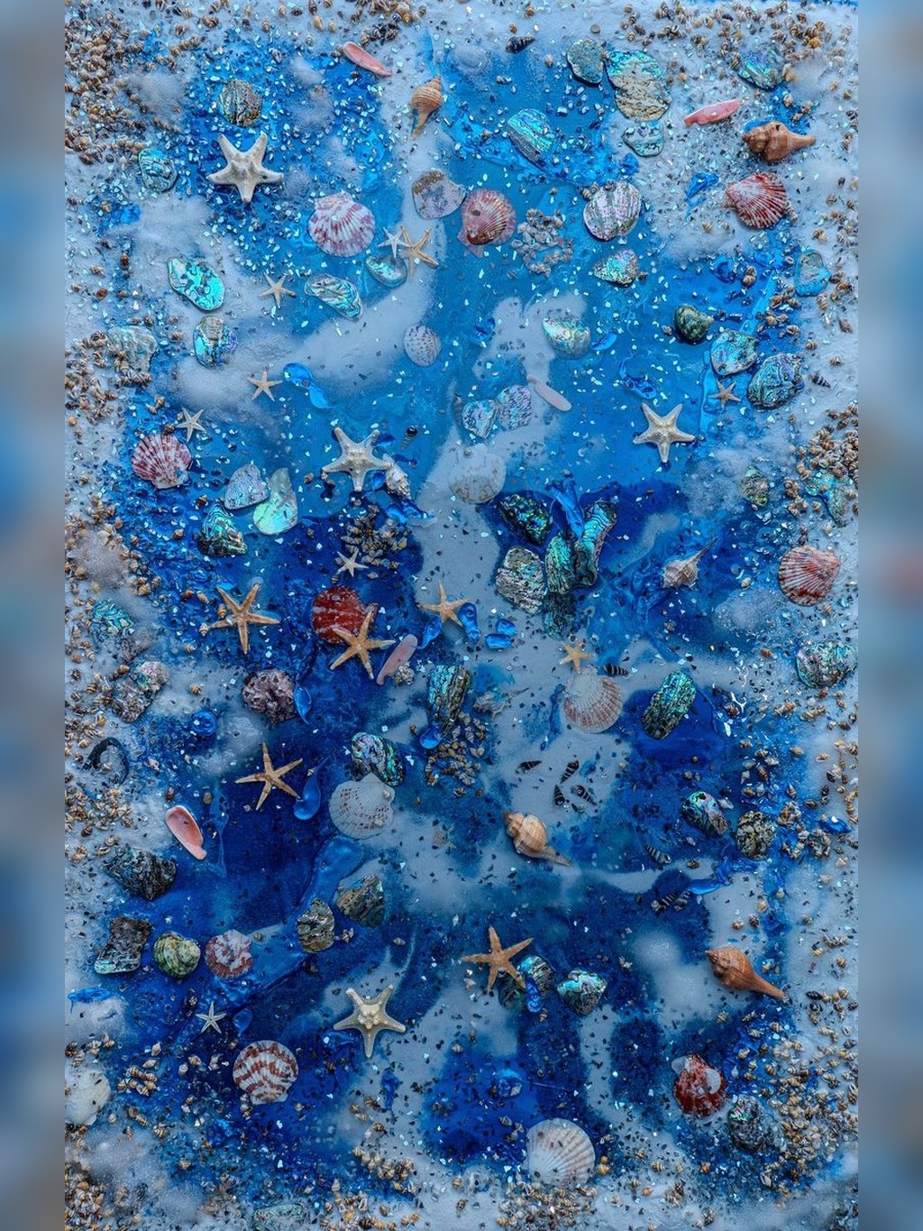 Blue-toned beach collage with assorted seashells, starfish, pebbles, and sand fragments scattered across a watery surface.
